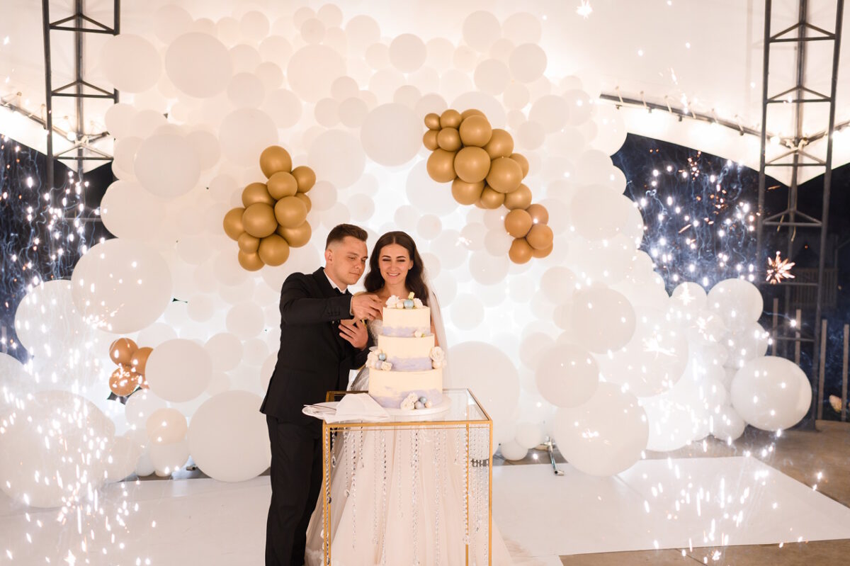Вид спереди красивых жениха и невесты, разрезая свадебный торт на фоне арки из воздушных шаров и фейерверков